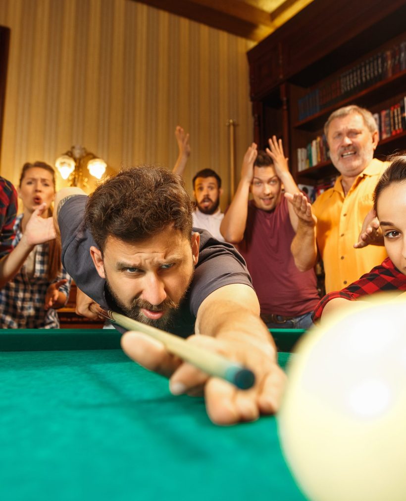 Close-up shot of a man playing billiard. The caucasian model carefully and strenuously aiming by cue in the ball. Game concept. Human emotions and facial expression concepts. Friends as background